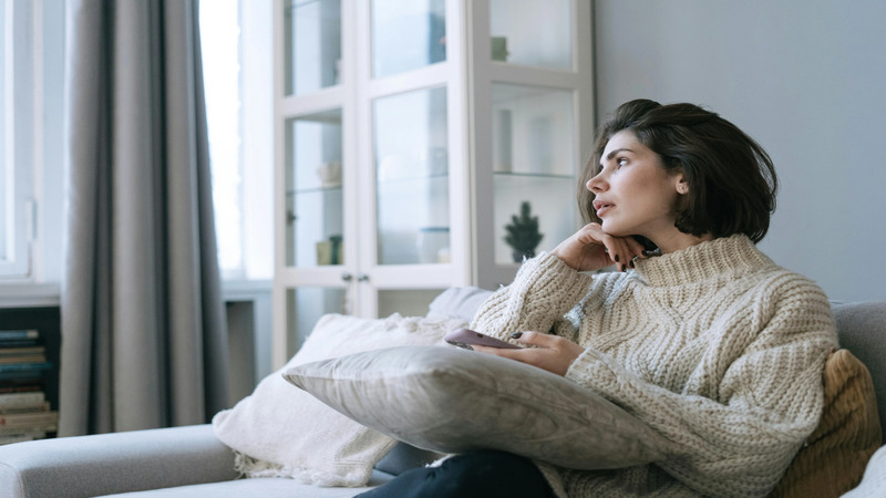 Person in a knitted sweater sitting on a couch, holding a pillow and looking to the side. A glass cabinet, large windows with curtains, and stacked books are in the background.